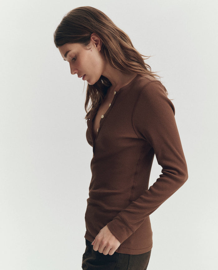 A person with long brown hair wears THE GREAT. PF26 RIB KNITS The Baby Rib Henley in Chocolate and dark pants, looking down while standing against a plain light background.