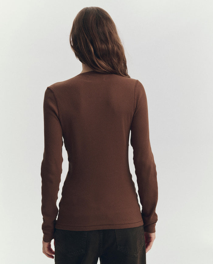 A person with long brown hair stands facing away, wearing THE GREAT. PF26 RIB KNITS The Baby Rib Henley in Chocolate and dark pants against a plain light background.