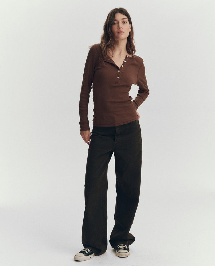 A person stands against a plain background, wearing THE GREAT. PF26 RIB KNITS The Baby Rib Henley in Chocolate, paired with dark wide-leg pants and black sneakers.
