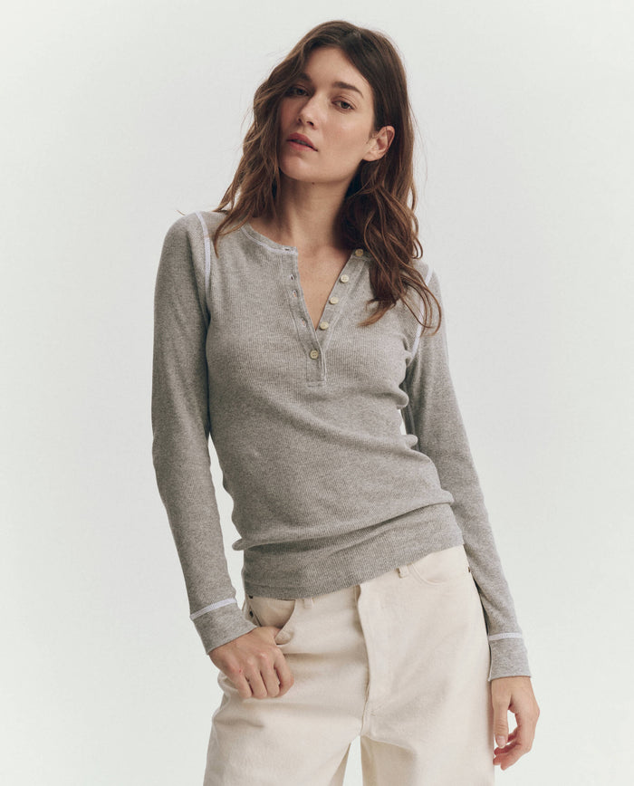 A woman wearing THE GREAT. F25 BABY RIB’s The Baby Rib Henley in Heather Grey with light-colored pants stands against a plain background.