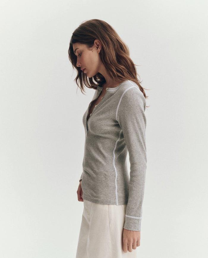 Woman with brown hair wears THE GREAT. F25 BABY RIB's The Baby Rib Henley in Heather Grey and cream pants, standing in profile against a plain light background.