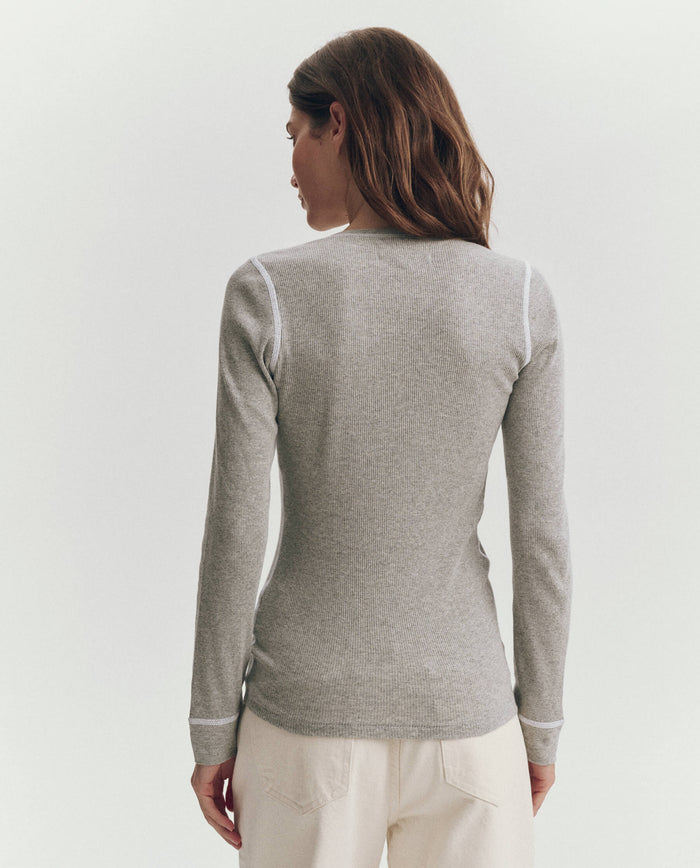 A woman faces away, wearing THE GREAT. F25 BABY RIB "The Baby Rib Henley" in heather grey with white trim and off-white pants, set against a plain background.