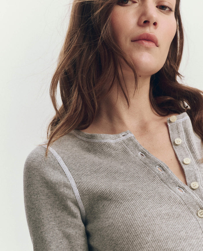 A woman with wavy brown hair models THE GREAT. F25 BABY RIB’s The Baby Rib Henley in Heather Grey, glancing away from the camera against a neutral background.