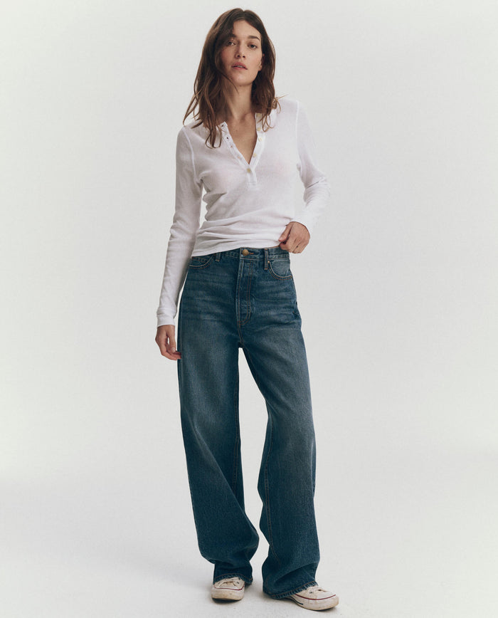 A person stands against a plain background wearing THE GREAT. F25 BABY RIB's The Baby Rib Henley in True White, loose blue recycled cotton jeans, and white sneakers.