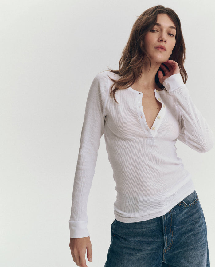 A person wearing THE GREAT. F25 BABY RIB The Baby Rib Henley in True White with blue jeans stands against a plain light background.