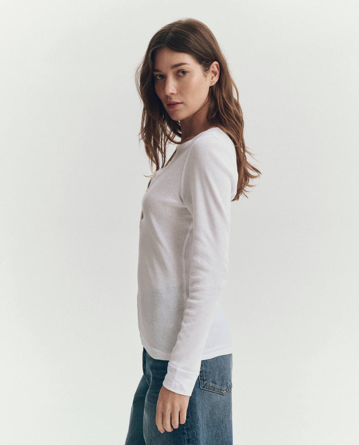 A person with long brown hair stands sideways against a plain white background, wearing THE GREAT. F25 BABY RIB The Baby Rib Henley in True White and blue jeans.