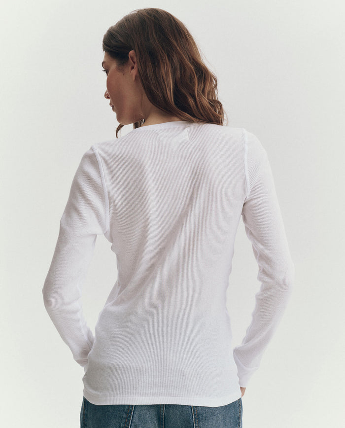 A woman with long brown hair faces away from the camera, wearing THE GREAT. F25 BABY RIB—The Baby Rib Henley in True White—and blue jeans against a plain background.