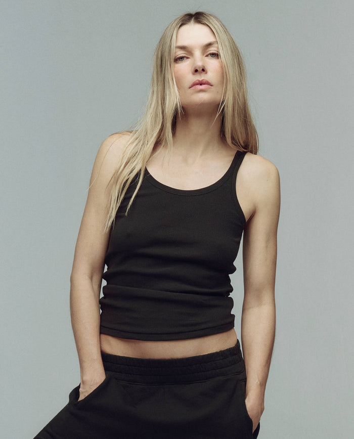 A person with long blonde hair wears THE GREAT. SU25 BABY RIB’s The Baby Rib Tank. -- Almost Black and black pants, standing against a plain gray background with hands in pockets.