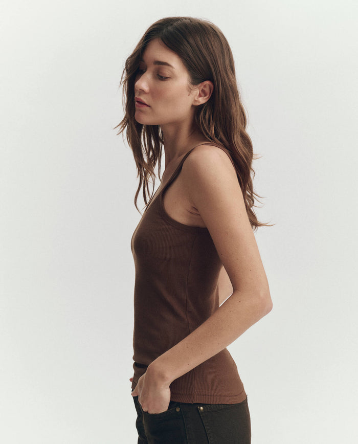 A woman with long brown hair stands in profile against a plain light background, wearing THE GREAT. PF26 RIB KNITS The Baby Rib Tank in Chocolate and dark pants.