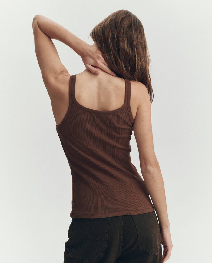 A person with long brown hair, seen from behind in dark pants, wears THE GREAT. PF26 RIB KNITS' The Baby Rib Tank in Chocolate, with a hand resting on their neck.