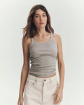 A woman with long brown hair wears THE GREAT. SU25 BABY RIB KNITS The Baby Rib Tank in Heather Grey and off-white pants against a plain light background. This vintage-inspired cotton tank is handcrafted in Los Angeles for timeless comfort.