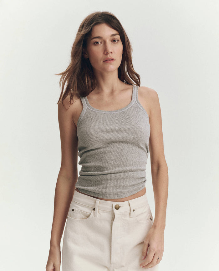 A woman with long brown hair wears THE GREAT. SU25 BABY RIB KNITS The Baby Rib Tank in Heather Grey and off-white pants against a plain light background. This vintage-inspired cotton tank is handcrafted in Los Angeles for timeless comfort.