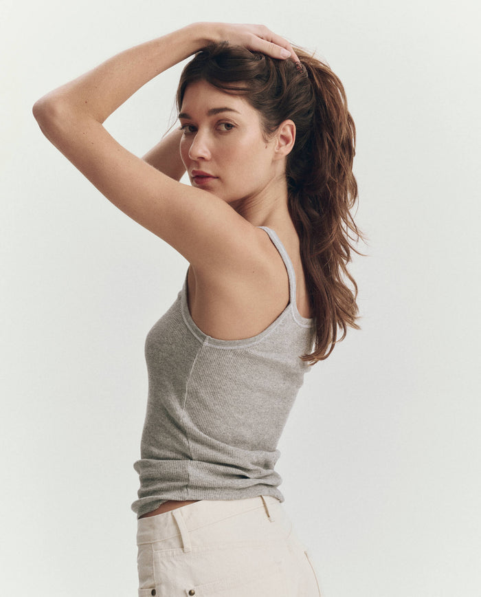 A woman wearing THE GREAT. SU25 BABY RIB KNITS The Baby Rib Tank in Heather Grey stands sideways against a plain light background, looking at the camera with one arm raised to hold her hair back.