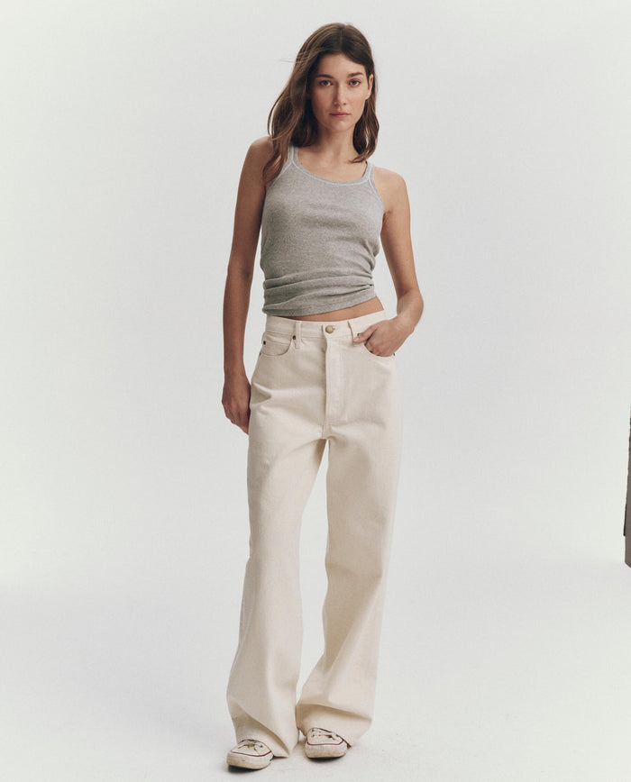 A woman poses against a plain background wearing THE GREAT. SU25 BABY RIB KNITS The Baby Rib Tank in Heather Grey, paired with cream wide-leg pants and white sneakers, one hand in her pocket.