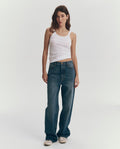 A woman stands against a plain background wearing THE GREAT. SU25 BABY RIB KNITS The Baby Rib Tank in True White, blue jeans, and white sneakers.