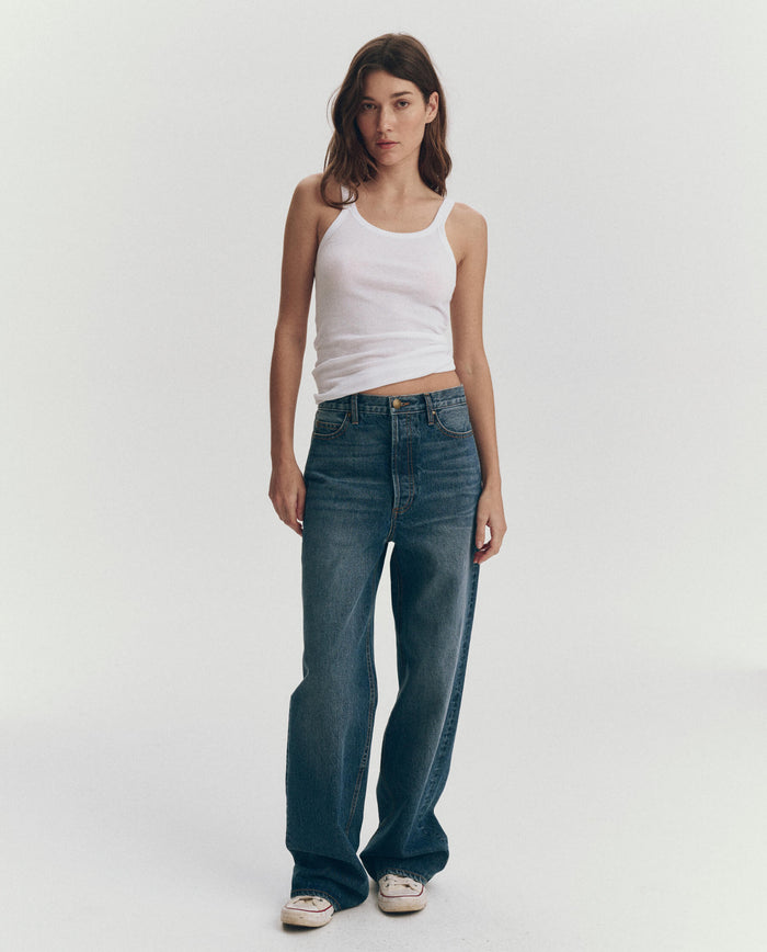 A woman stands against a plain background wearing THE GREAT. SU25 BABY RIB KNITS The Baby Rib Tank in True White, blue jeans, and white sneakers.
