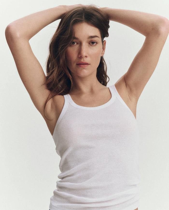 A woman wears THE GREAT.'s The Baby Rib Tank in True White, posing with arms raised and hands on her head, looking at the camera against a plain background.