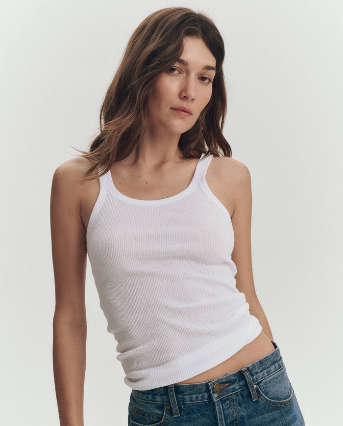 A woman wears THE GREAT. SU25 BABY RIB KNITS The Baby Rib Tank in True White with blue jeans, standing against a plain light background, looking at the camera with a neutral expression.