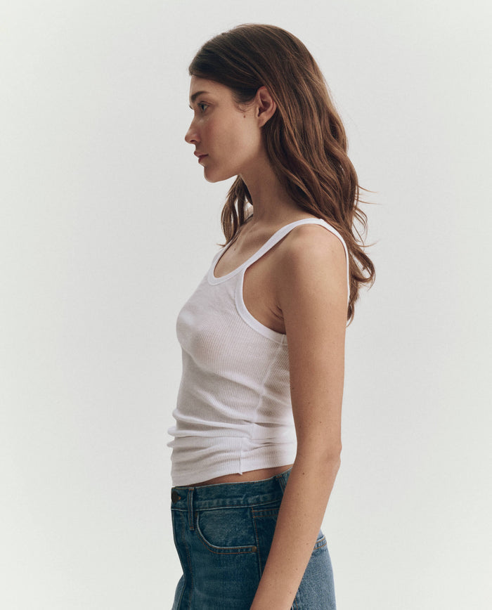A woman stands in profile against a light background, wearing THE GREAT. SU25 BABY RIB KNITS "The Baby Rib Tank" in True White styled as a classic cotton tank with blue jeans.