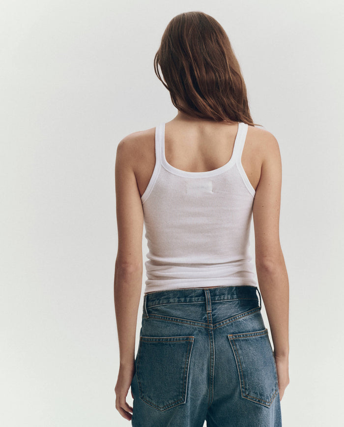 Someone faces away from the camera, wearing THE GREAT. SU25 BABY RIB KNITS "The Baby Rib Tank" in True White and blue jeans, set against a plain light background.