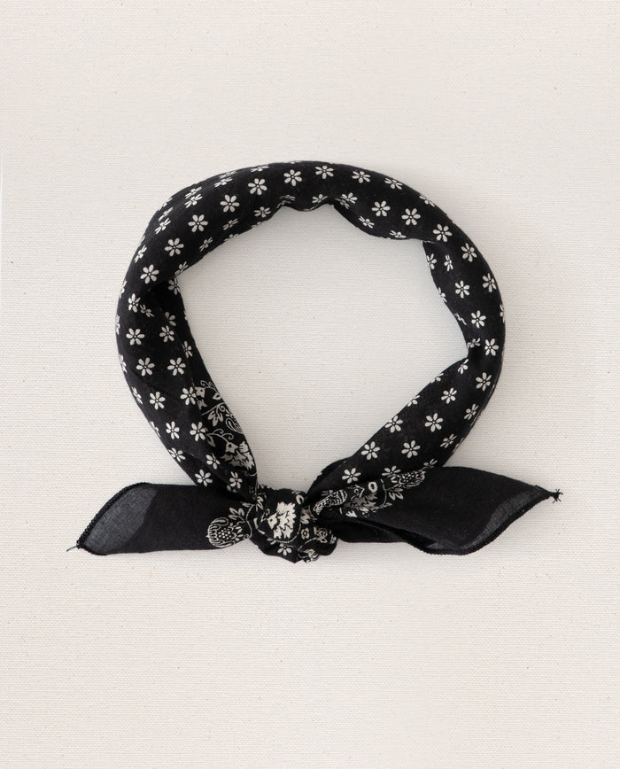 A small "Almost Black" bandana from THE GREAT. SP24 D2, adorned with white floral and paisley patterns, folded and tied in a simple knot, reminiscent of vintage handkerchiefs. Crafted for an ultra-soft feel with 100% cotton.