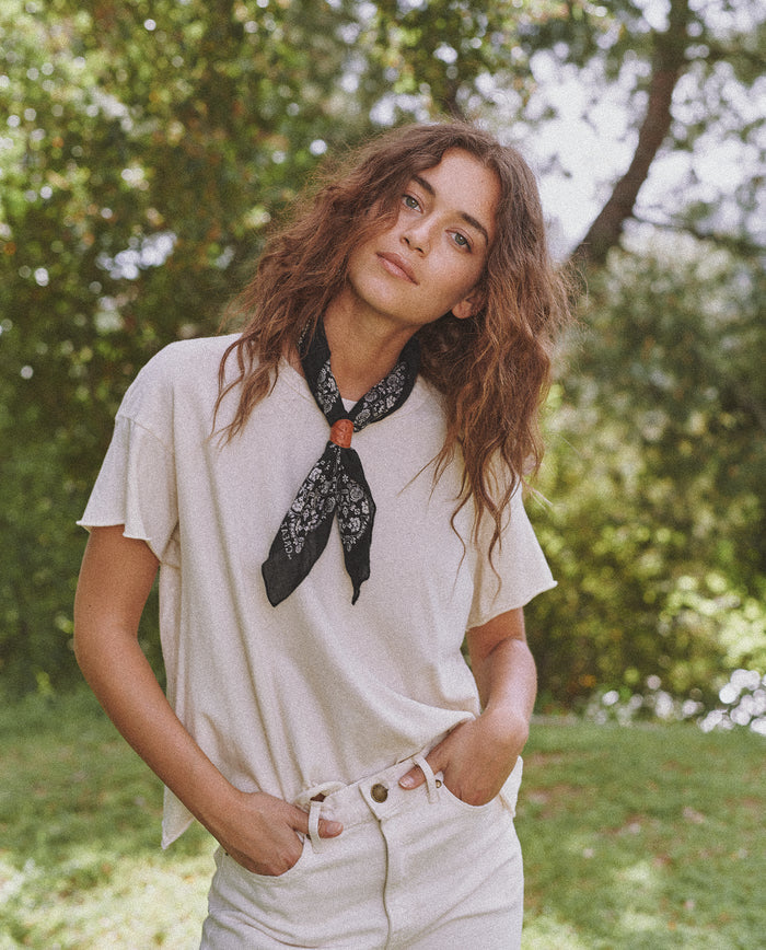 A person with curly hair wearing a white t-shirt, white pants, and "The Bandana - Small" in Almost Black by THE GREAT. SP24 D2 stands outdoors with greenery in the background.