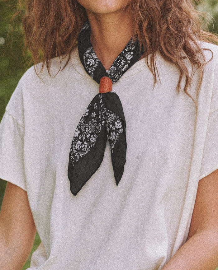 A person with long hair wearing a white t-shirt and The Bandana. -- Small -- Almost Black from THE GREAT. SP24 D2 around their neck, reminiscent of vintage handkerchiefs.
