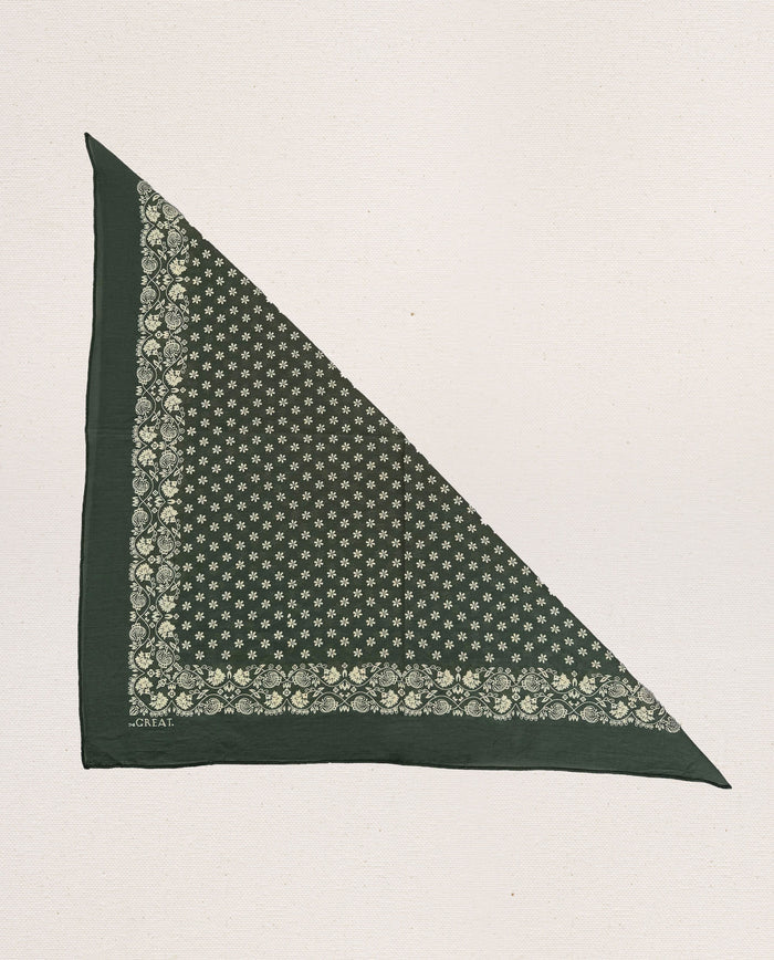 The Bandana – Small in Army Green, from THE GREAT. SP24 D2, is a meticulously crafted piece made from 100% cotton. This green triangular bandana showcases an intricate white floral and geometric design along its borders and inner area. Set against a plain background, it embodies the charm of vintage handkerchiefs while delivering an ultra-soft feel.