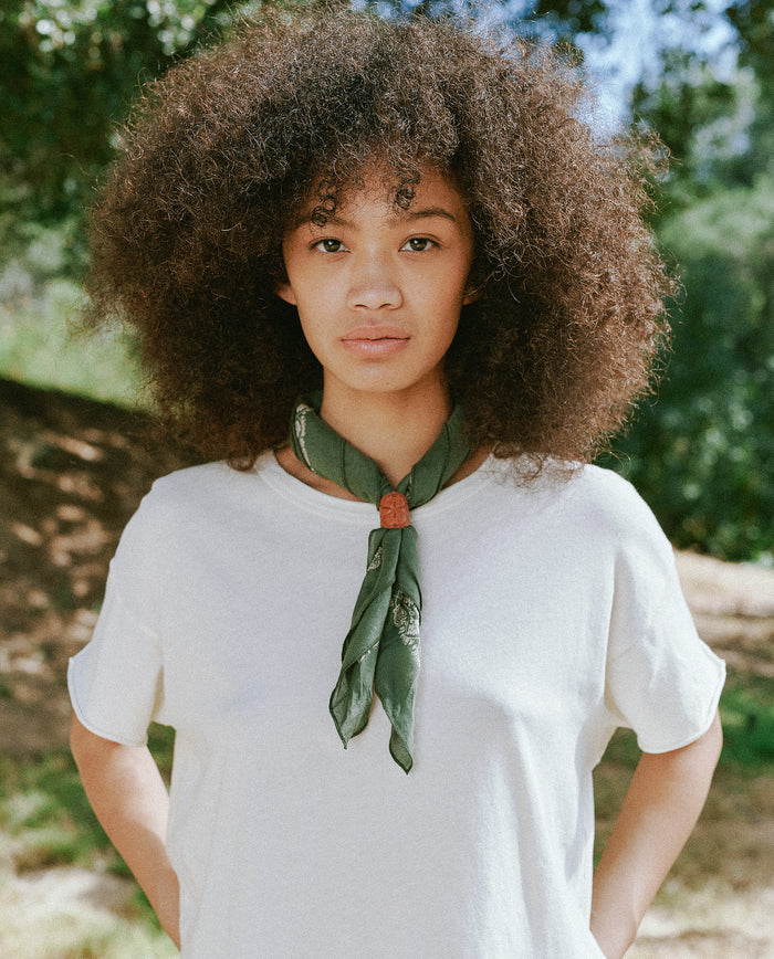 A person with curly hair stands outdoors wearing a white shirt and The Bandana in Army Green, made of ultra-soft hand 100% cotton from THE GREAT. SP24 D2. Trees are visible in the background.