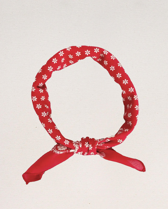 The Bandana – Small in Bright Red by THE GREAT. CARRYOVER, crafted from ultra-soft cotton, showcases white floral patterns on a vibrant red base and is tied in a knot against a plain, light background.
