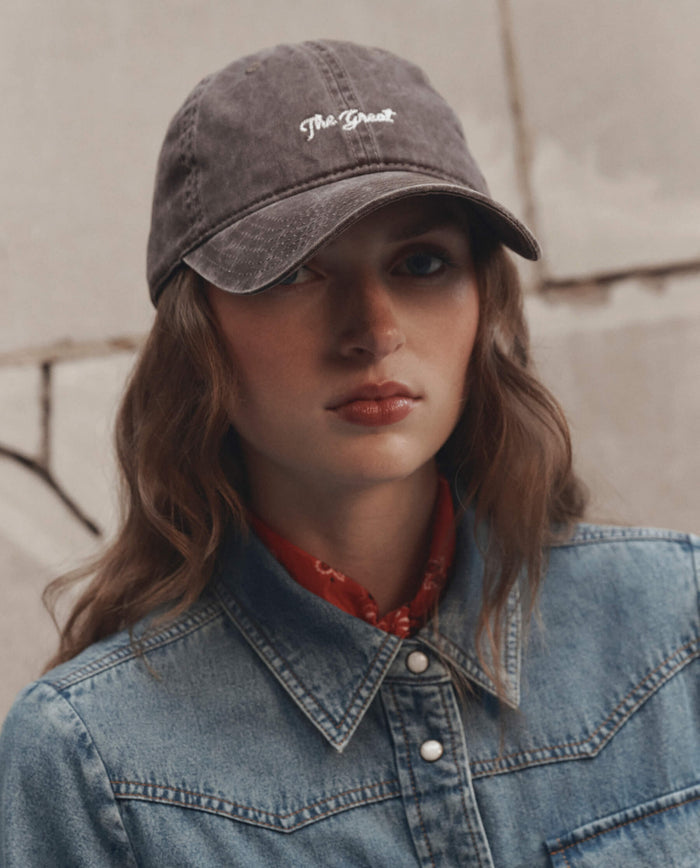 A person wearing The Baseball Hat in Chocolate by THE GREAT. F26 D1, a red scarf, and a blue denim jacket, standing against a concrete wall.