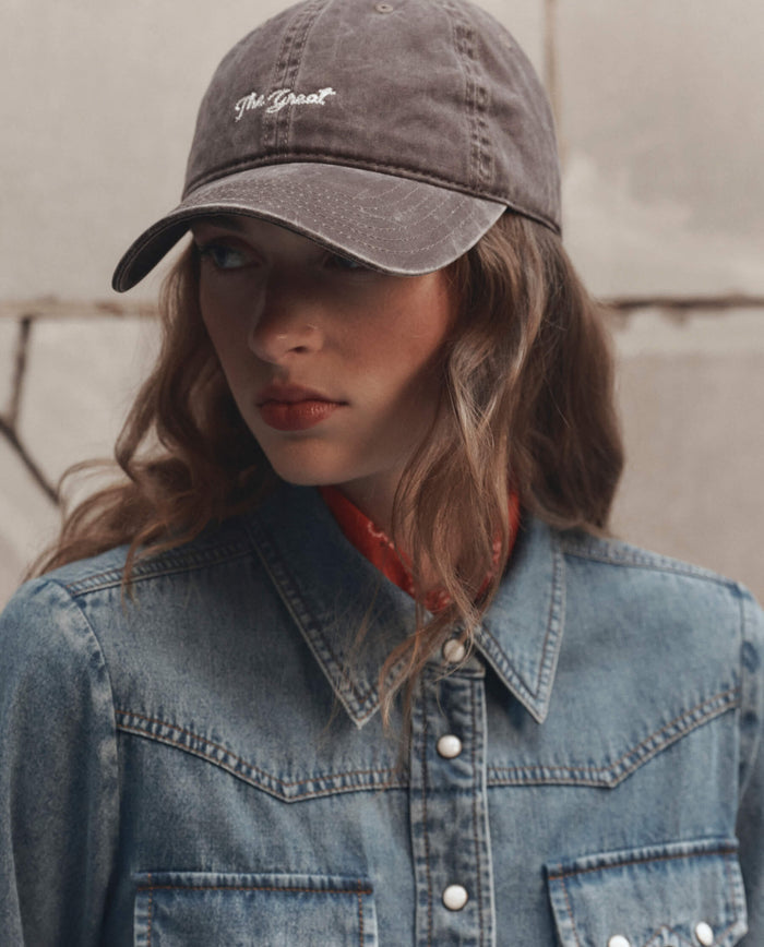 Woman in a denim shirt and The Baseball Hat in Chocolate by THE GREAT. F26 D1, looking to the side against a stone wall background.