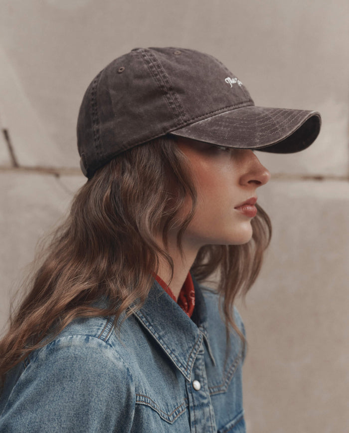 Wearing THE GREAT. F26 D1 The Baseball Hat in Chocolate and a denim jacket, a person stands in profile against a neutral stone background.