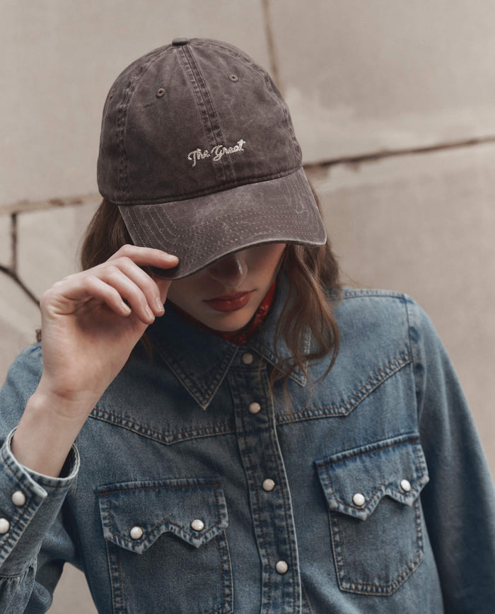 A person wearing a denim shirt and THE GREAT. F26 D1 The Baseball Hat in Chocolate, with "The Great" embroidered on the faded black cap, partially obscuring their face as they hold the brim.