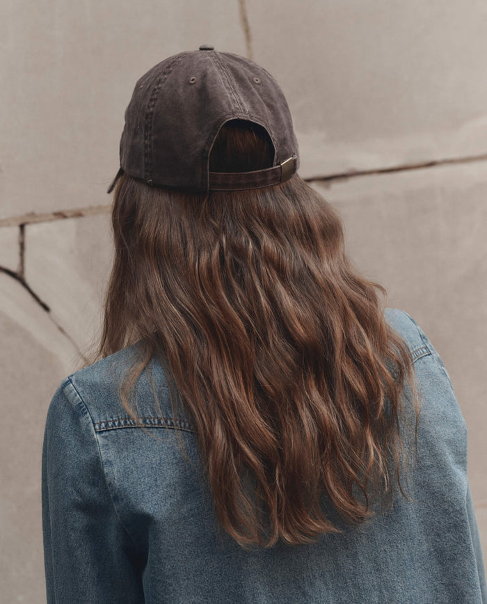 A person with long wavy brown hair, wearing THE GREAT. F26 D1 The Baseball Hat in Chocolate and a blue denim jacket, faces away from the camera against a light-colored wall.