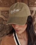 A person wearing THE GREAT. PF26 "The Baseball Hat" in khaki, with the brand's name embroidered on the front, looking down.