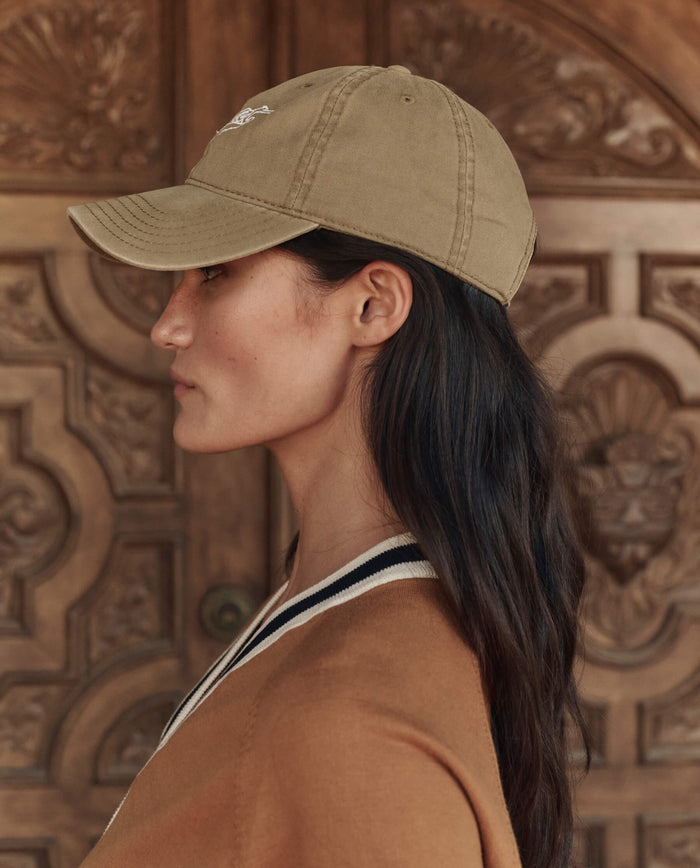 A person with long dark hair stands in profile before a carved wooden door, wearing THE GREAT. PF26 "The Baseball Hat" in khaki and a tan jacket with black and white trim.
