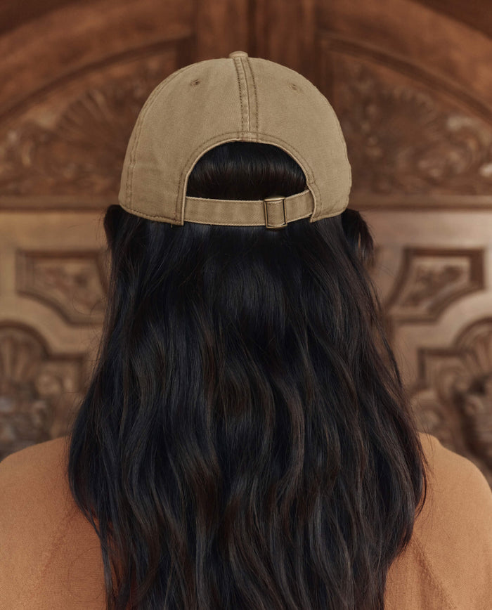 A person with long dark hair, seen from behind, wears THE GREAT. PF26 The Baseball Hat in Khaki while standing in front of a carved wooden door.