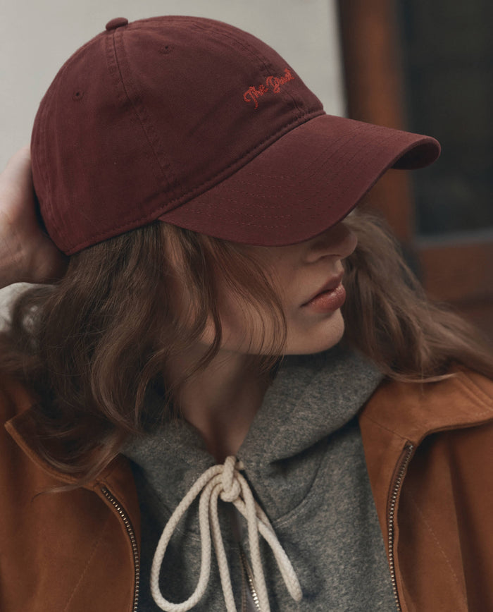 Wearing THE GREAT. F26 D1 The Baseball Hat in Maroon, a person dressed in a brown jacket and grey hoodie with white drawstrings looks to the side, adjusting their cap.
