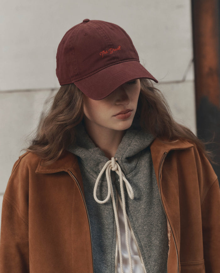 A person with long hair wears THE GREAT. F26 D1 "The Baseball Hat." in maroon, a grey hoodie with a white drawstring, and a brown suede jacket against a textured wall.
