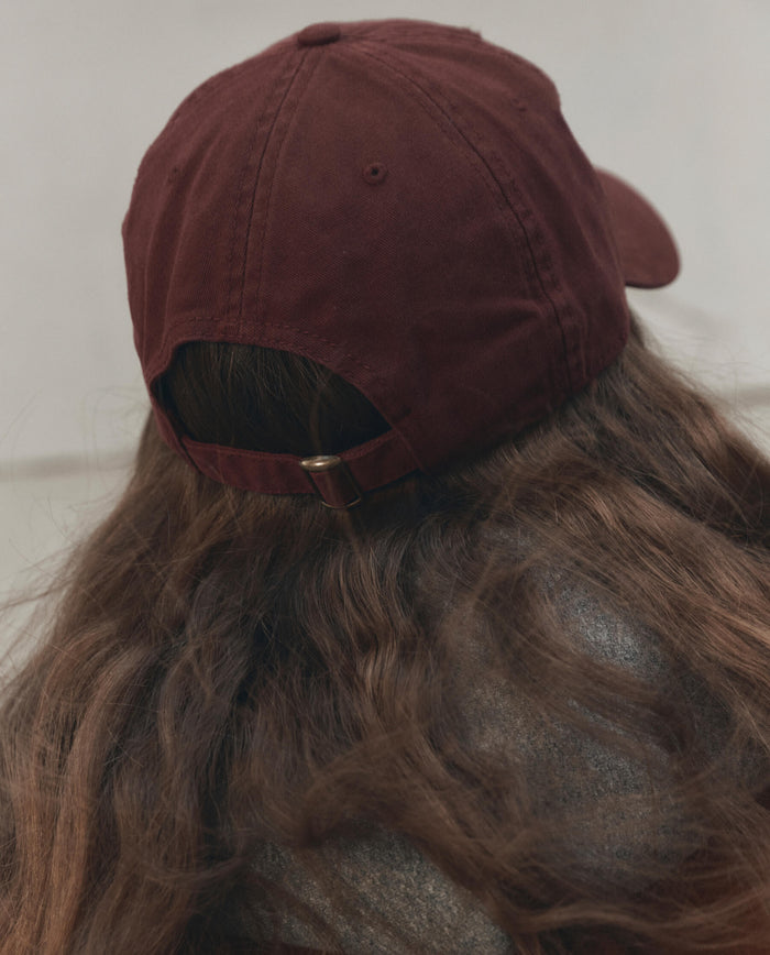 Person with long brown hair, photographed from behind against a plain background, wearing THE GREAT. F26 D1 The Baseball Hat in maroon.