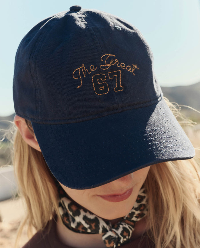 A person wearing THE GREAT. PF26 The Baseball Hat in navy and a leopard print scarf around their neck.