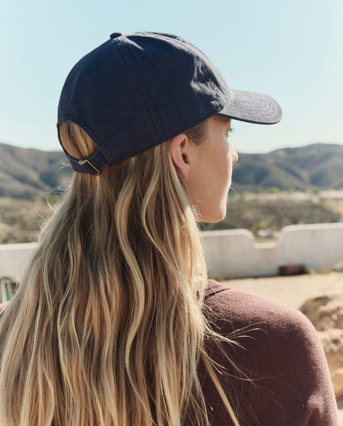 A person with long blonde hair is outdoors, facing away from the camera, wearing THE GREAT. PF26 The Baseball Hat in navy and a brown shirt, with hills and a clear sky visible in the background.