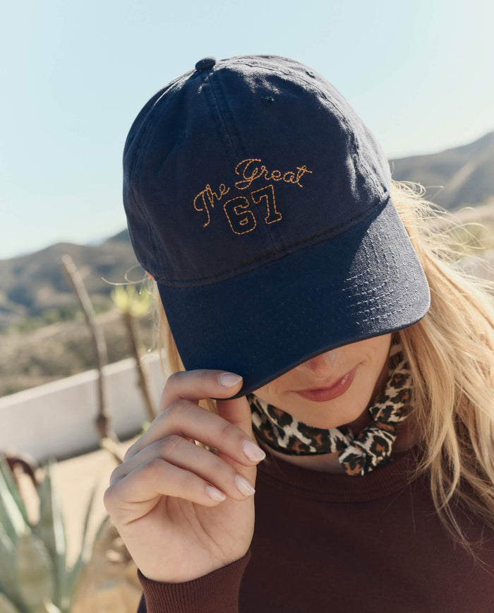 A person wearing THE GREAT. PF26 The Baseball Hat in navy, featuring "The Great 67" embroidered in yellow, stands outdoors near desert plants and mountains.