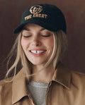 A woman wearing THE GREAT. F25 D1 ACCESSORIES The Baseball Hat. Graphic -- Black, featuring "THE GREAT" in chainstitch embroidery, smiles while looking slightly down, paired with a tan jacket and gray top.