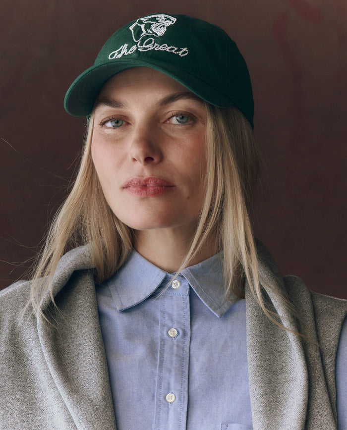 A woman with long blonde hair and a neutral expression wears THE GREAT. F25 D1 ACCESSORIES The Baseball Hat. Graphic -- Green, featuring vintage "The Great" and bear embroidery, paired with a light blue button-up shirt and gray sweater.