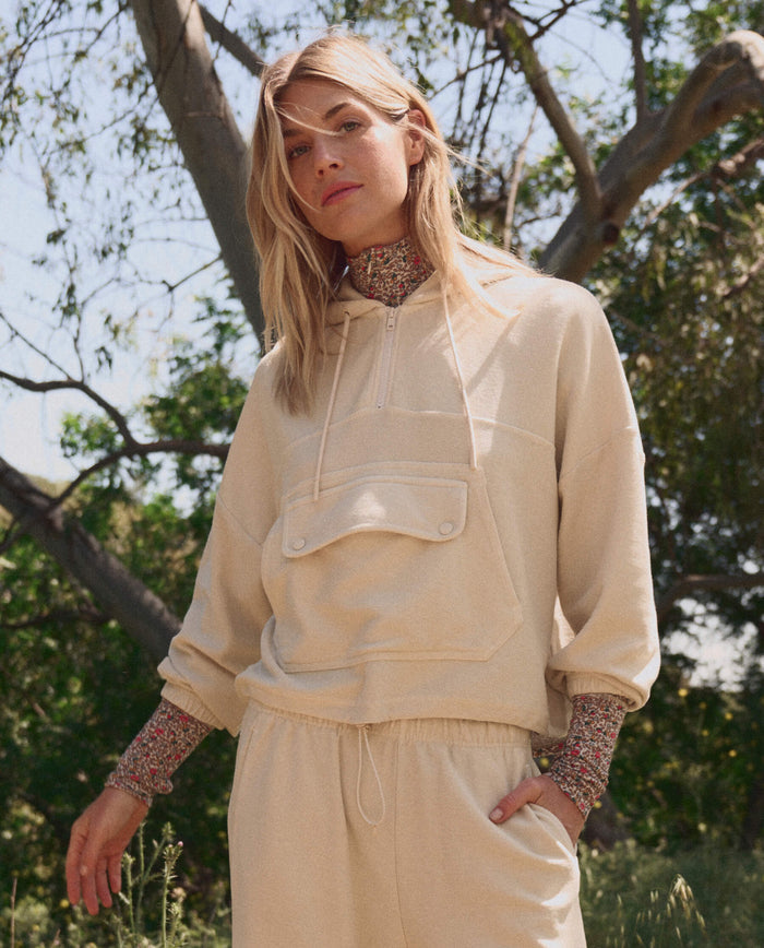 A woman stands outdoors in THE GREAT. SP26 TGO’s The Basecamp Pullover—Oatmilk, a beige hoodie with front pocket and matching pants—layered over a floral turtleneck, perfect for outdoor moments among trees and greenery.