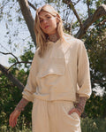 A woman stands outdoors in THE GREAT. SP26 TGO’s The Basecamp Pullover—Oatmilk, a beige hoodie with front pocket and matching pants—layered over a floral turtleneck, perfect for outdoor moments among trees and greenery.