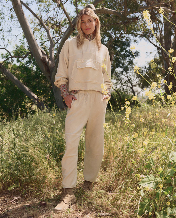 A woman stands outdoors among tall grass and wildflowers, wearing THE GREAT. SP26 TGO’s The Basecamp Pullover in Oatmilk—ideal outdoor apparel—with trees and sunlight in the background.