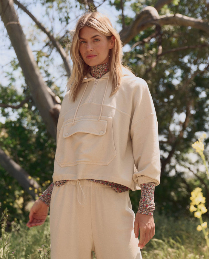 A woman enjoys the outdoors in THE GREAT. SP26 TGO The Basecamp Pullover — Oatmilk, styled with matching pants and a floral turtleneck, framed by trees and sunlight for a chic outdoor look.